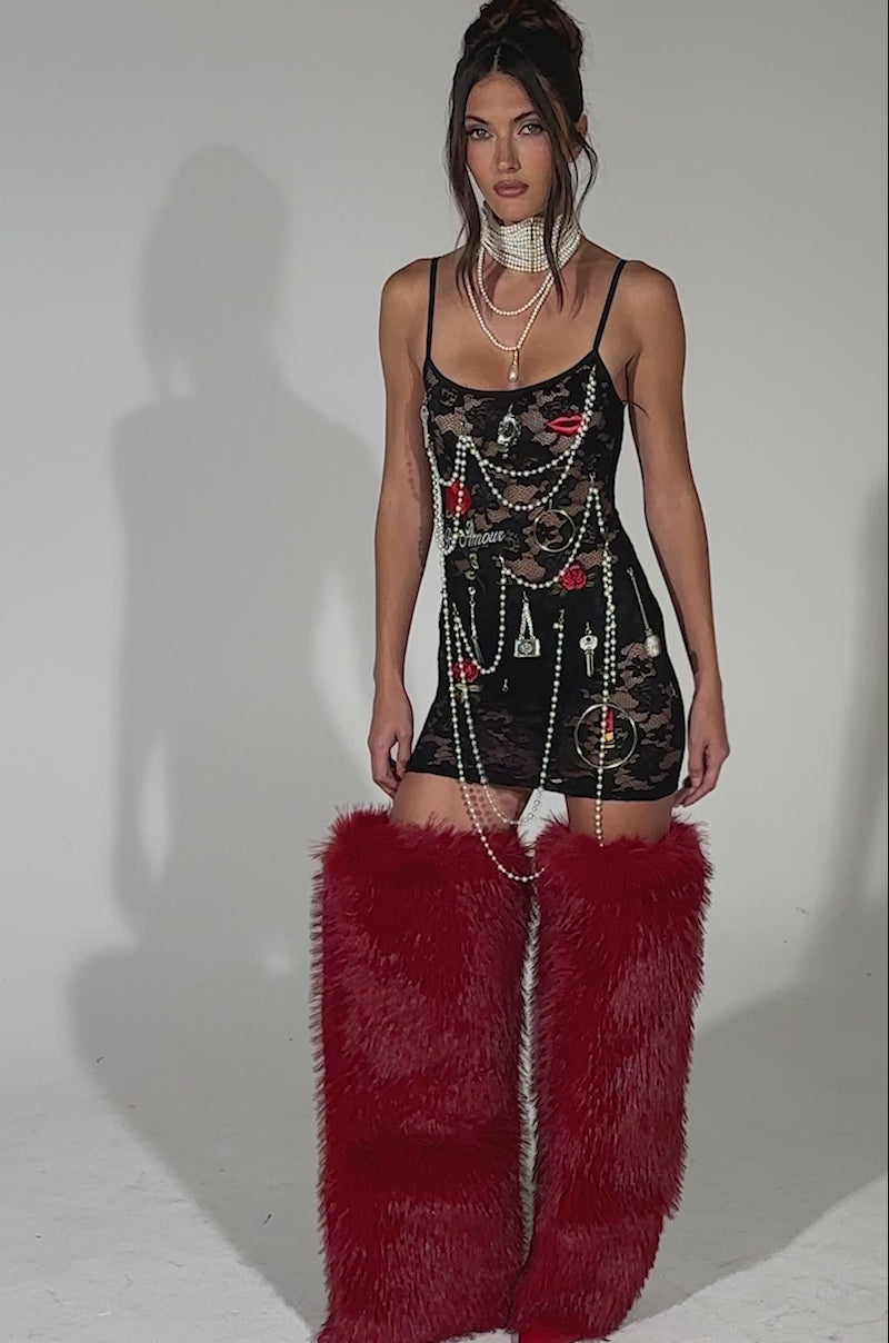 A video of a woman posing in the FEELING CRAZY SEAMLESS JUNK MINI DRESS—a short black pearl chain dress with decorative charms—layered pearl necklaces, and bold tall red furry boots against a plain light background.