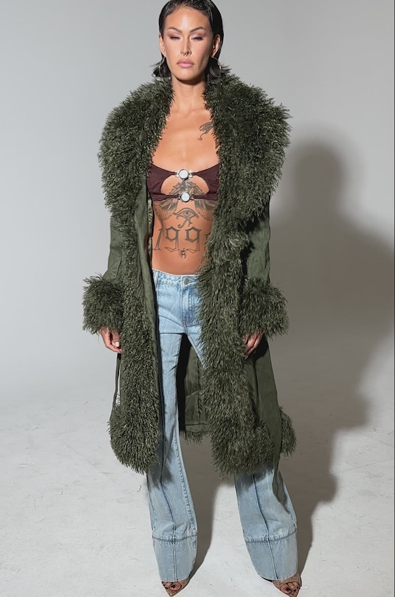 Video of woman modeling the JADE FUR TRIM TRENCH in olive green, featuring thick faux fur trim on the collar, cuffs, and hem, belted at the waist and styled over blue jeans against a plain background.