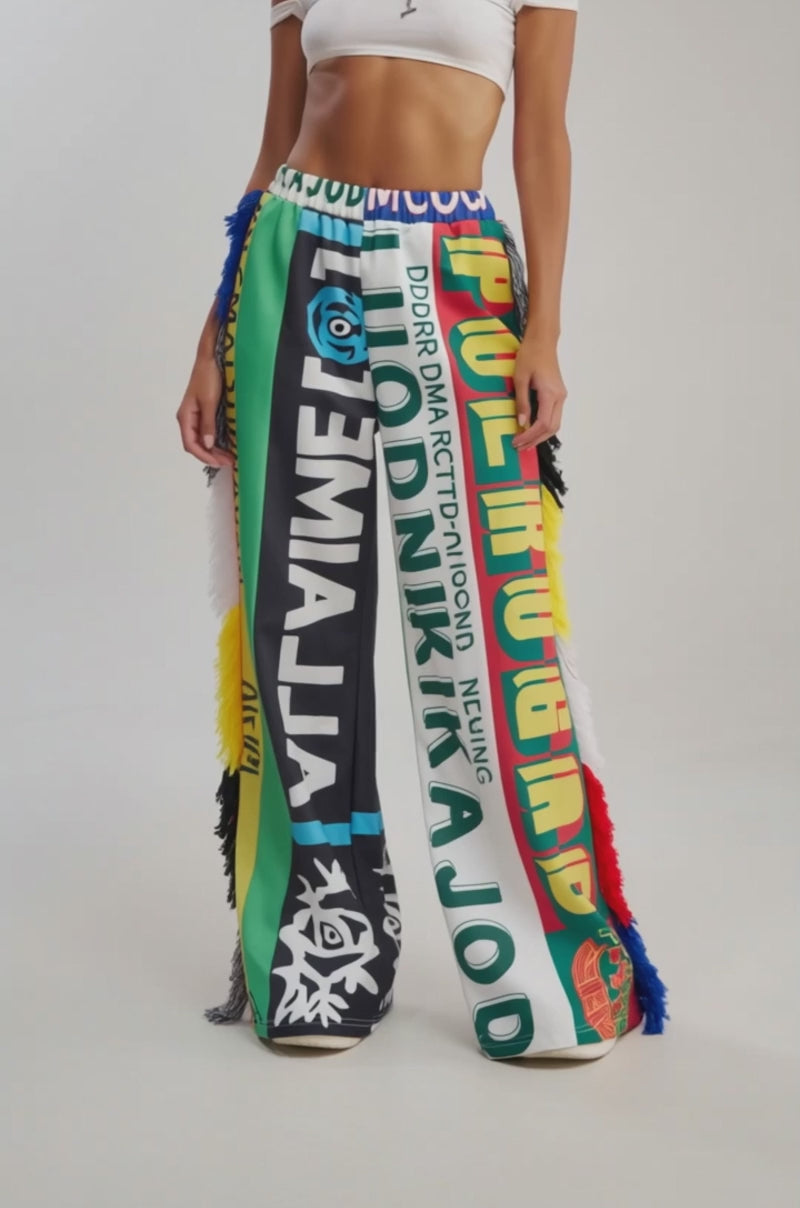 Video of woman modeling the MIXED EMOTIONS GRAPHIC JOGGER, featuring colorful wide-leg pants with bold graphic patterns, multicolored text, and fringed sides, paired with white shoes. 