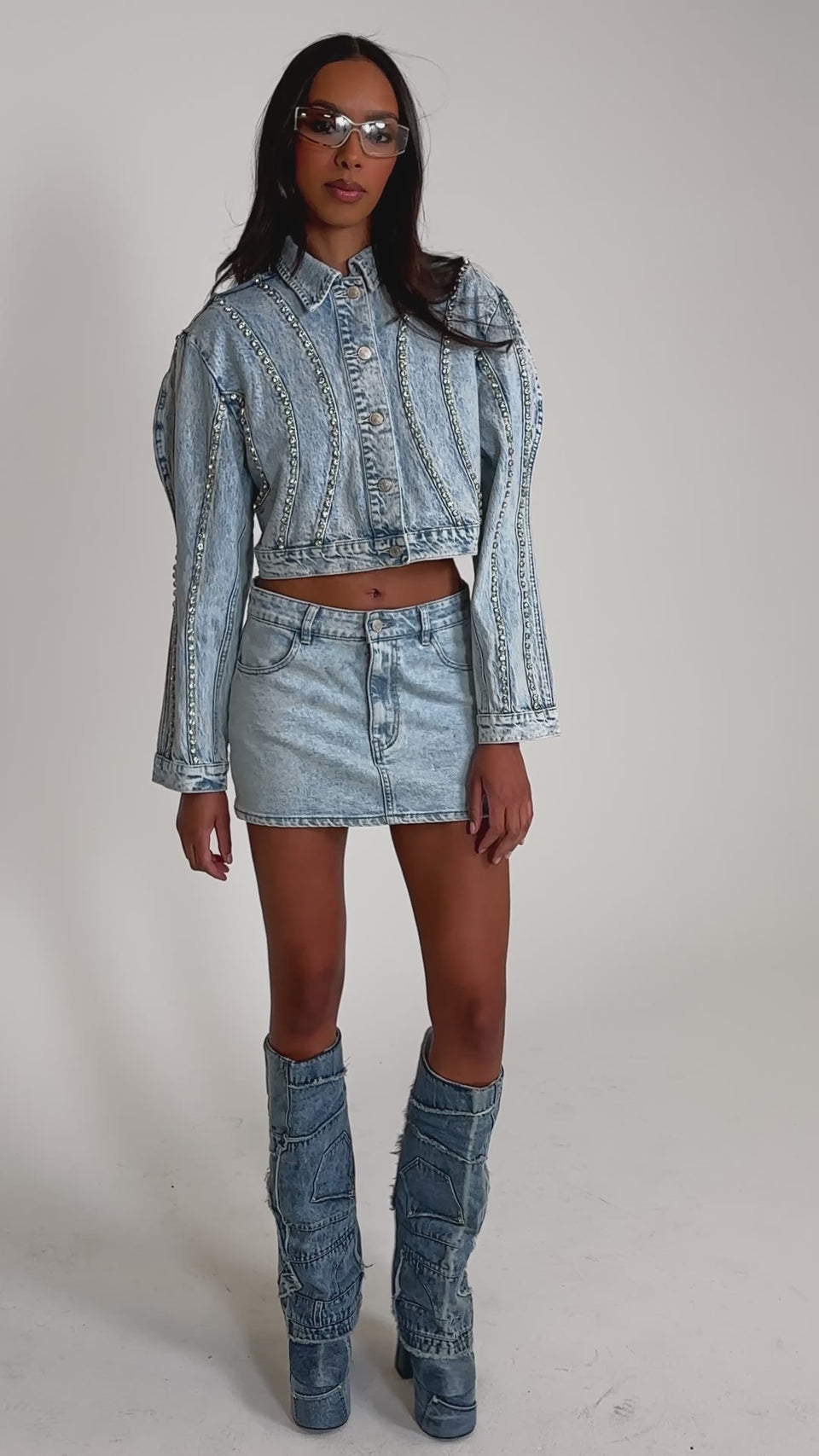 Video of woman modeling the EXTRA EXTRA EMBELLISHED DENIM JACKET IN LIGHT BLUE, paired with a matching denim skirt and clear-frame glasses, posing confidently against a simple background.
