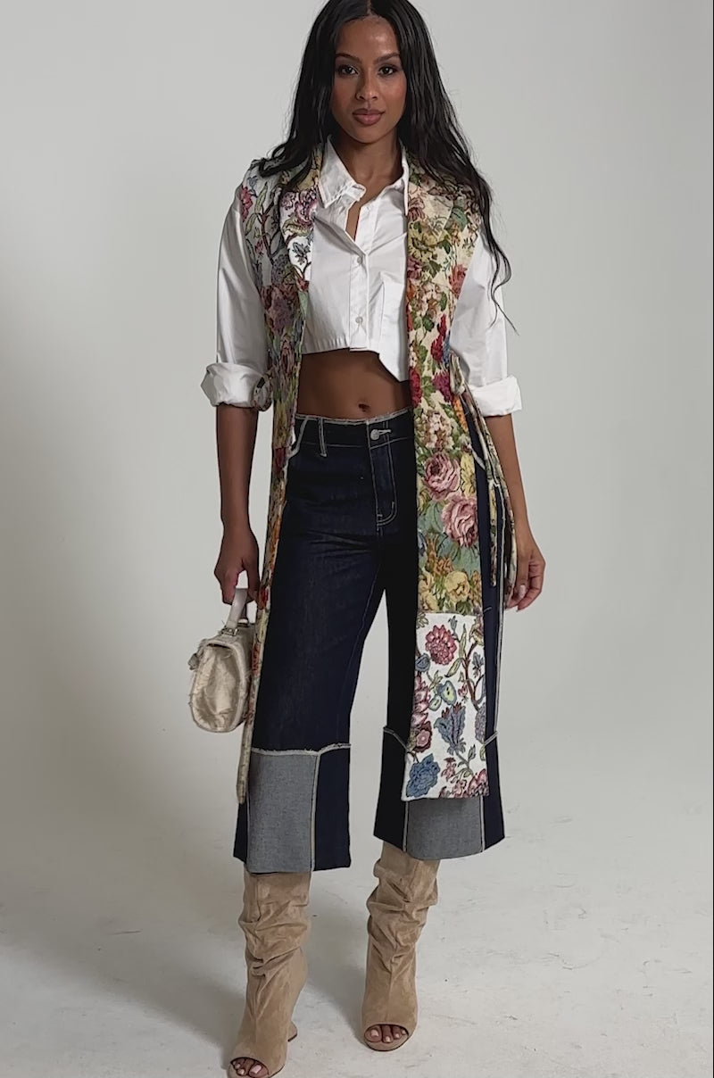 Video of woman modeling the HONEY HONEY SPLIT VEST IN TAPESTRY over a white cropped shirt, high-waisted dark cuffed jeans, and tan knee-high boots.