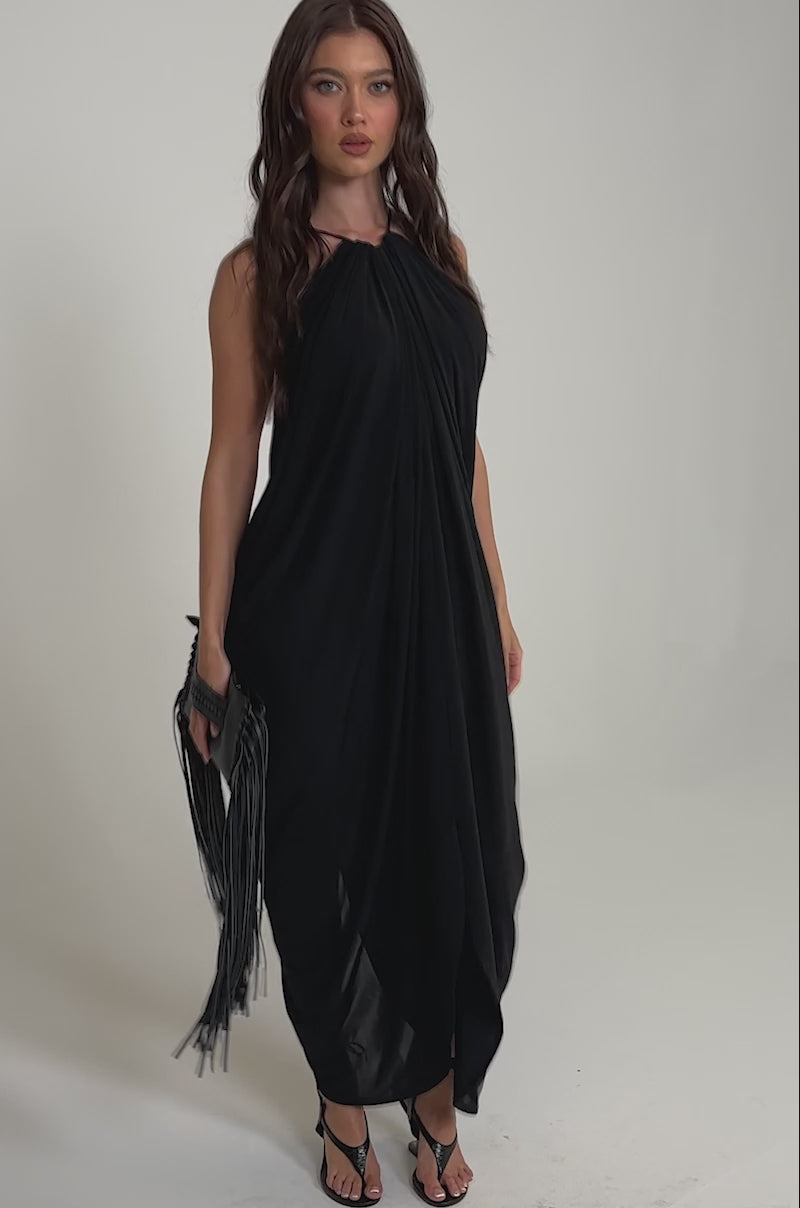 A video of a woman in the BALI MAMI LOW BACK MAXI DRESS IN BLACK, featuring a loose drape, gathered neckline, and low-cut back. 