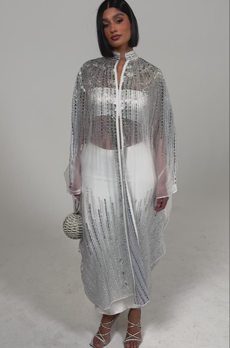 Video of woman modeling the SPARKLE AND SHINE BLOUSE IN WHITE under a sheer, silver-sequined cape dress, paired with a round silver handbag and strappy silver heels, standing against a plain light background.