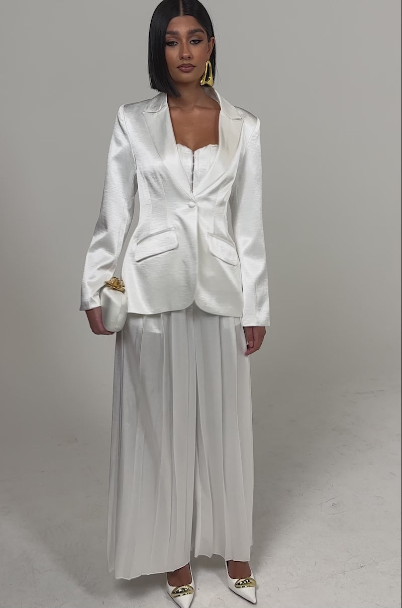 A video of a woman in the IRIS SATIN BLAZER, tailored at the waist and styled with a long pleated skirt, gold pointed heels, bold gold earrings, and sleek straight hair.