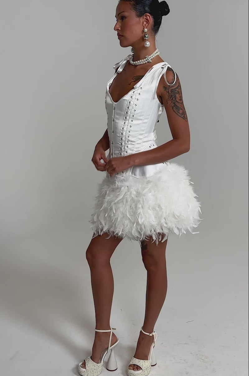 A video of a woman in the LITTLE FAIRY FEATHER MINI SKIRT with a white lace-up corset top, platform heels, and large earrings.