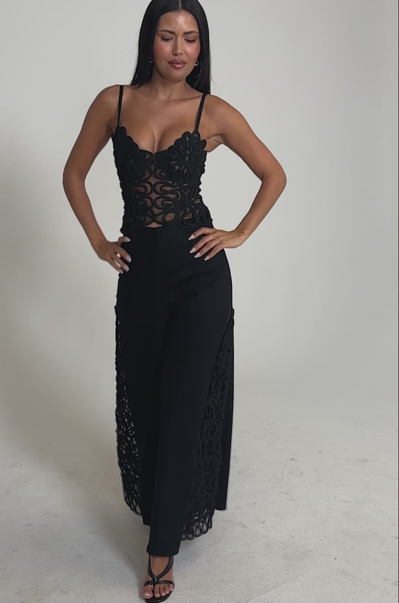 Video of woman modeling the ITS GIVING ROPE DETAIL TROUSER, a pair of high-waisted, wide-leg black pants with intricate rope and sheer cut-out details, styled with strappy black heels against a plain background.