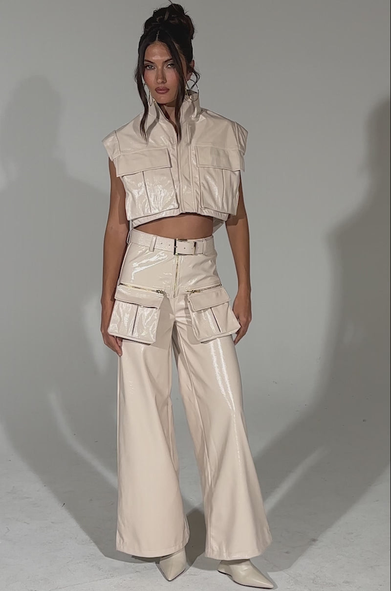A video of a woman posing against a light background, wearing SHOW STOPPER FAUX LEATHER CARGO TROUSER—a shiny, sleeveless light beige cropped jacket with front pockets and matching high-rise, wide-leg cargo trousers with oversized pockets and pointed-toe shoes.