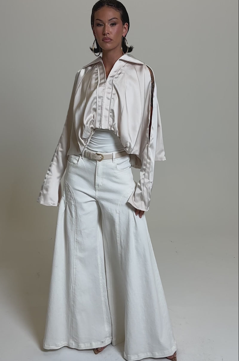 A video of a person in the GET WITH IT SATIN BLOUSE—light beige with Dolman sleeves—paired with high-waisted, wide-leg white pants, a belt, and large hoop earrings against a plain grey background.