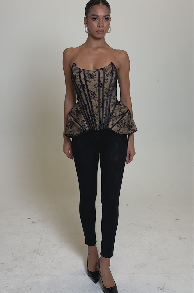 A video of a woman wearing the JUST A GIRL LACE CORSET, a strapless black floral piece, paired with black pants, hoop earrings, pointed black heels, and styled hair pulled back, all set against a plain light background.