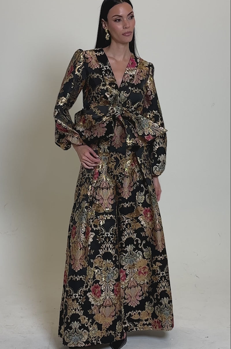 A video of a woman in the Luxe Energy Palazzo Pant, featuring a black brocade floral print with gold, pink, and green accents. She poses against a neutral backdrop, highlighting the pant's striking wide-leg silhouette and peplum jacket.