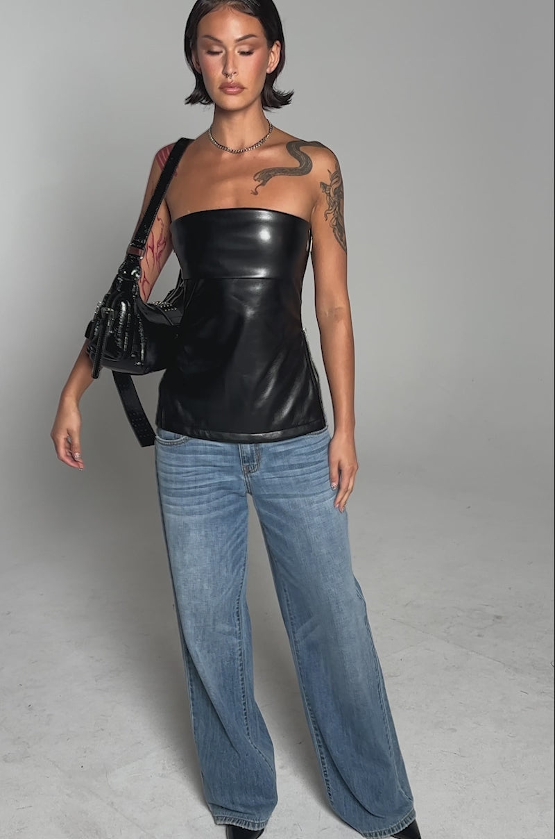 A video of a person with short dark hair and arm tattoos wearing the ZIP ME UP ON THE SIDE CORSET in black faux leather, styled with loose blue jeans, pointed shoes, and a black shoulder handbag.