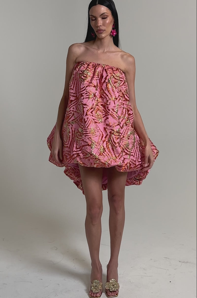 A video of a woman wearing the LOVER GIRL BROCADE BUBBLE DRESS IN PINK, a strapless mini dress with a pink and gold metallic finish, voluminous silhouette, and high-low hem. 