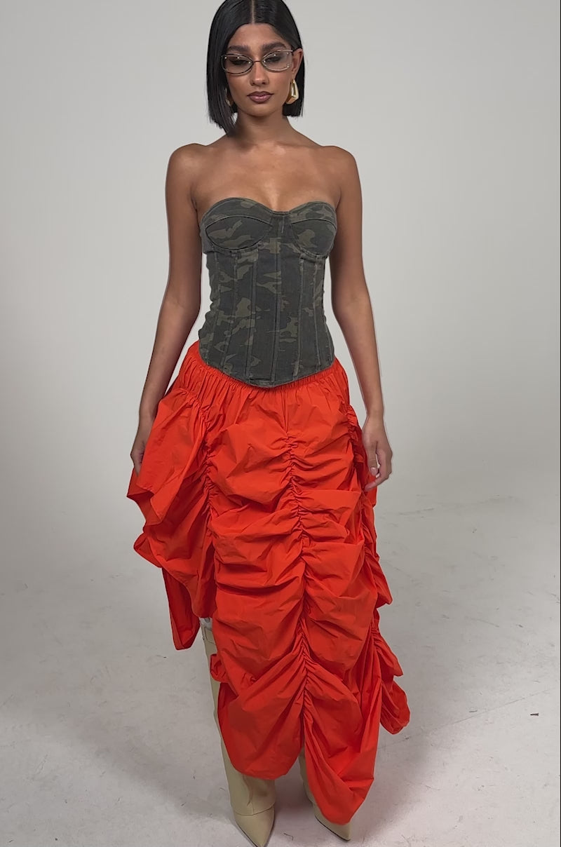 A video of a woman in a strapless camouflage corset top, the LETS CHANGE IT UP RUCHED MIDI SKIRT IN ORANGE, beige knee-high boots, and clear glasses against a plain light background.