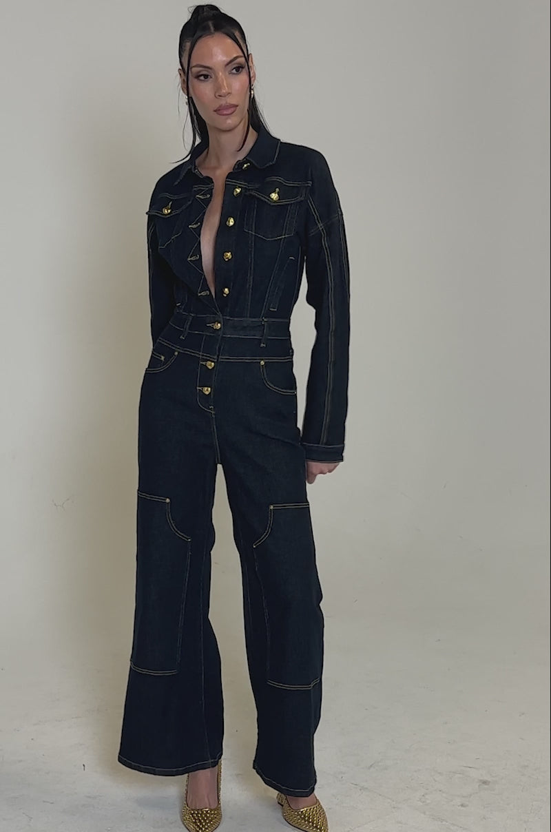 A video of a woman standing confidently against a plain, light background wearing the ANGELA CARGO DENIM JUMPSUIT—a dark blue, long-sleeved utility design with gold buttons, wide legs, and paired with gold heels.