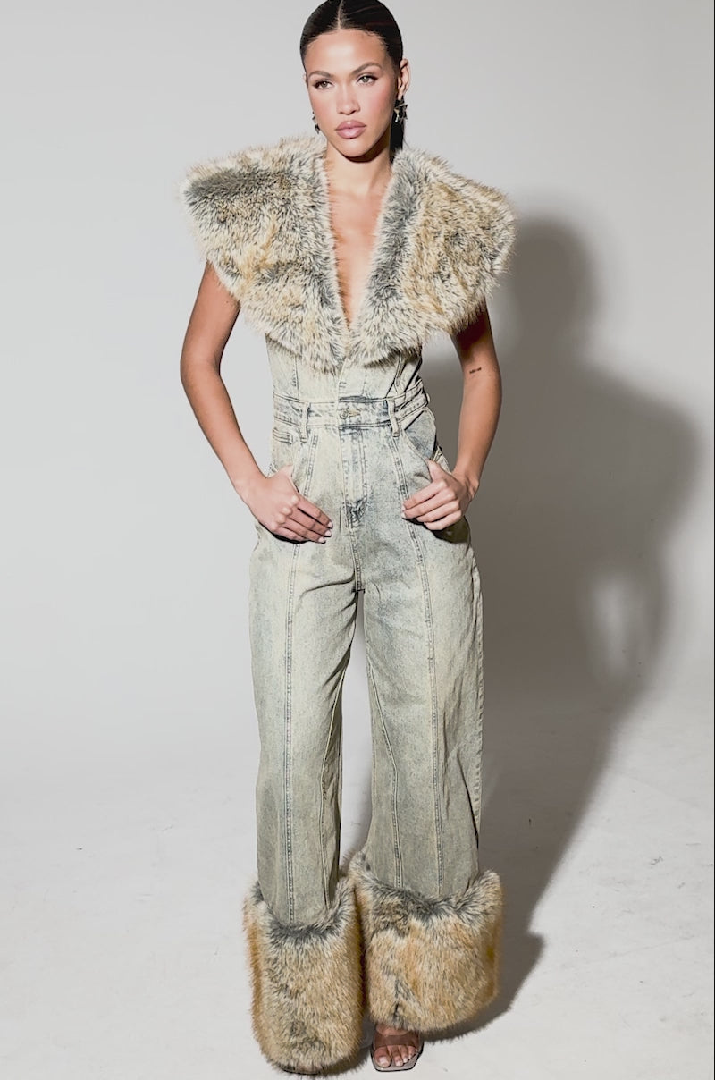 Video of woman modeling the TALK THAT TALK DENIM FUR JUMPSUIT IN VINTAGE BLUE, a deep V-neck, wide-leg denim piece trimmed with tan faux fur at the hems. Her hair is tied back and she pairs it with high-heeled sandals.