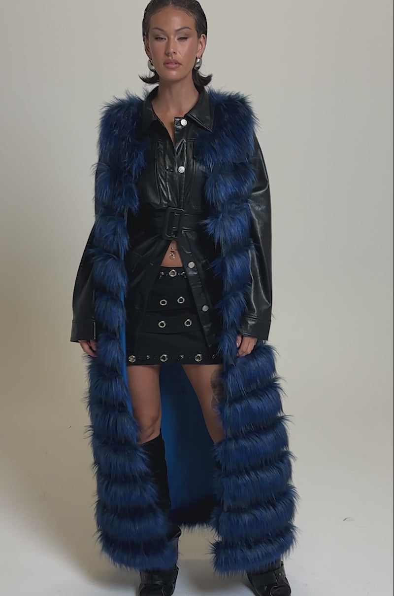 A video of a woman wearing the RIZZA PANELED FUR VEST in blue and black over a black belted jacket and short skirt with metal eyelets, styled with knee-high black boots.