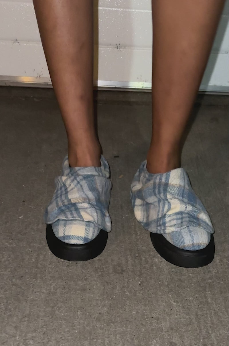 A video of a model posing on concrete in front of a white garage door, a person wearing ripped light blue denim shorts and AZALEA WANG SEYMON BLUE MULTI PLAID SLIP-ON RUCHED FLATS—with a treaded rubber sole—shows only their legs.