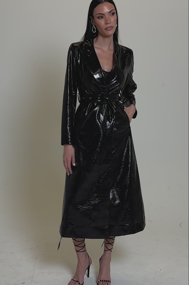 A video of a wearing the ACQUIRED TASTE FAUX LEATHER TRENCH featuring a glossy finish and belted waist, paired with strappy heels and hoop earrings against a neutral background.