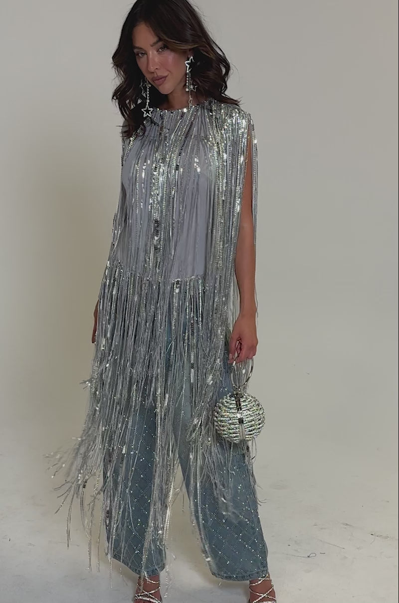 Video of woman modeling the DRIP CITY FRINGE TOP, a silver sequin statement piece with long metallic tassels, styled over wide-leg blue jeans. She completes her party look with a round woven handbag and silver high-heeled sandals.