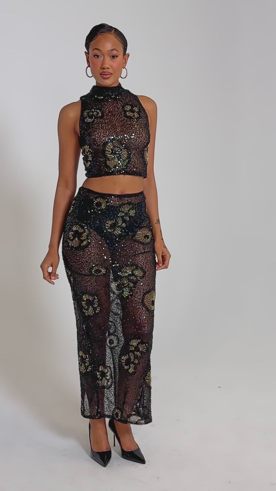A video of a woman wearing the ALL EYES ON ME FLORAL MOCKNECK TOP, a black sheer sequin crop top with floral patterns, paired with a matching skirt, hoop earrings, slicked-back hair, and black pointed heels.
