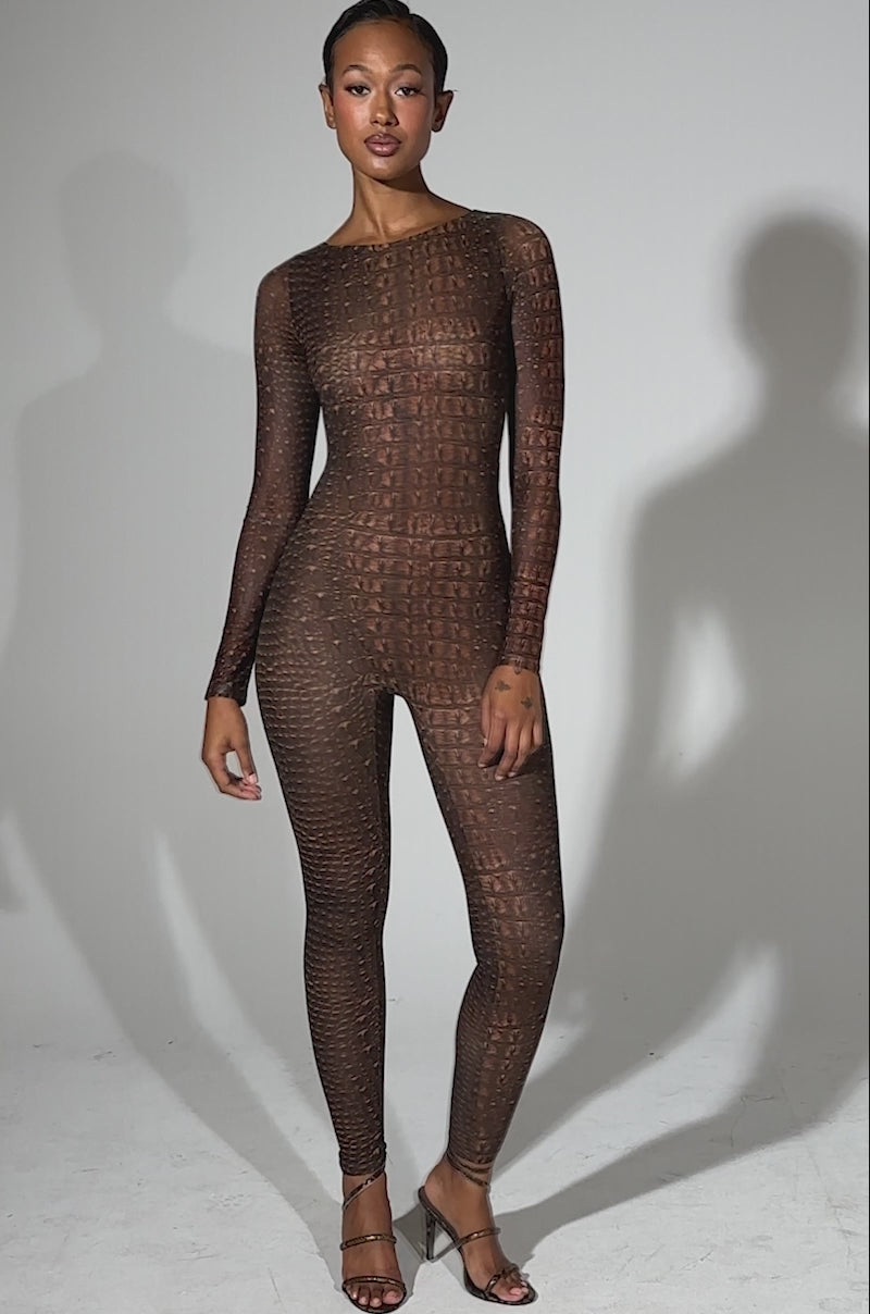A video of a woman posing confidently in the TRY AND GET ME SNAKE MESH JUMPSUIT, featuring a brown snake print, mesh fabric, long sleeves, and black strappy heels against a plain light gray background.