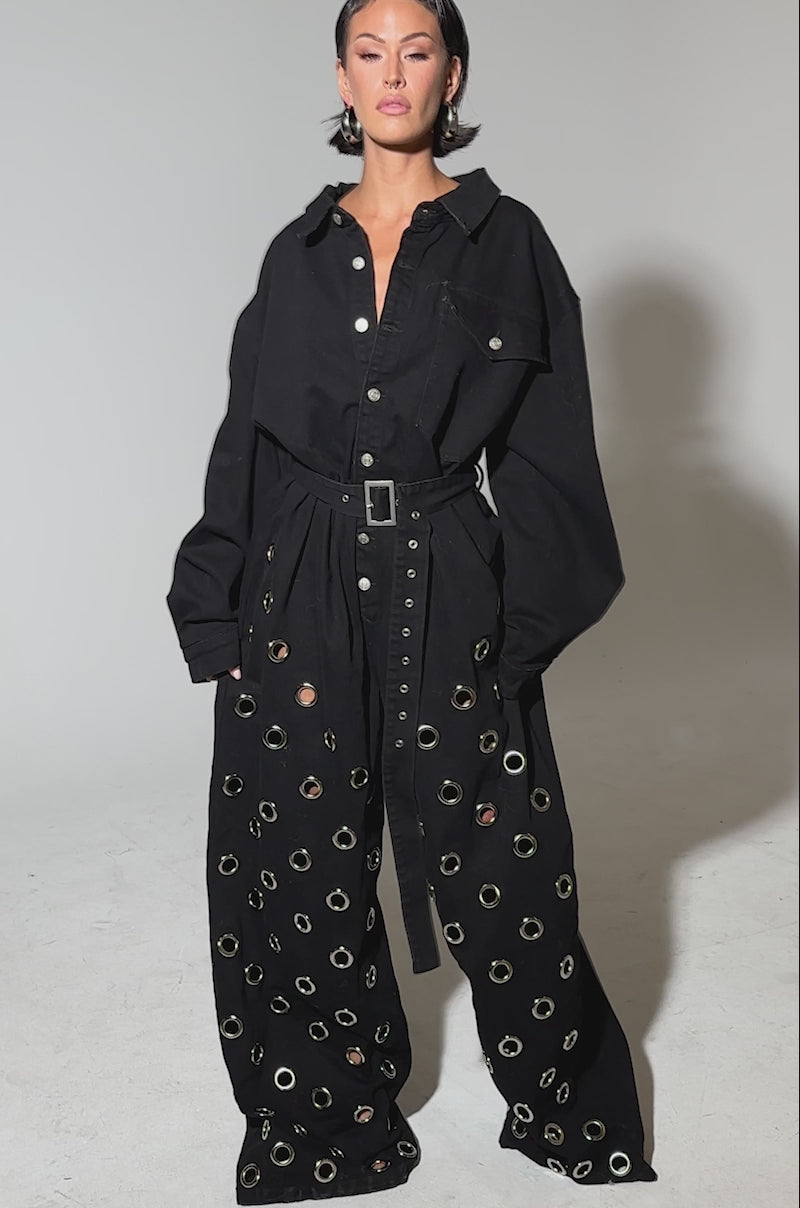 Video of woman modeling the PLAY YOUR CARDS RIGHT DENIM JUMPSUIT WITH GROMMETS, featuring a loose fit, belted waist, silver grommet details, and wide legs. 