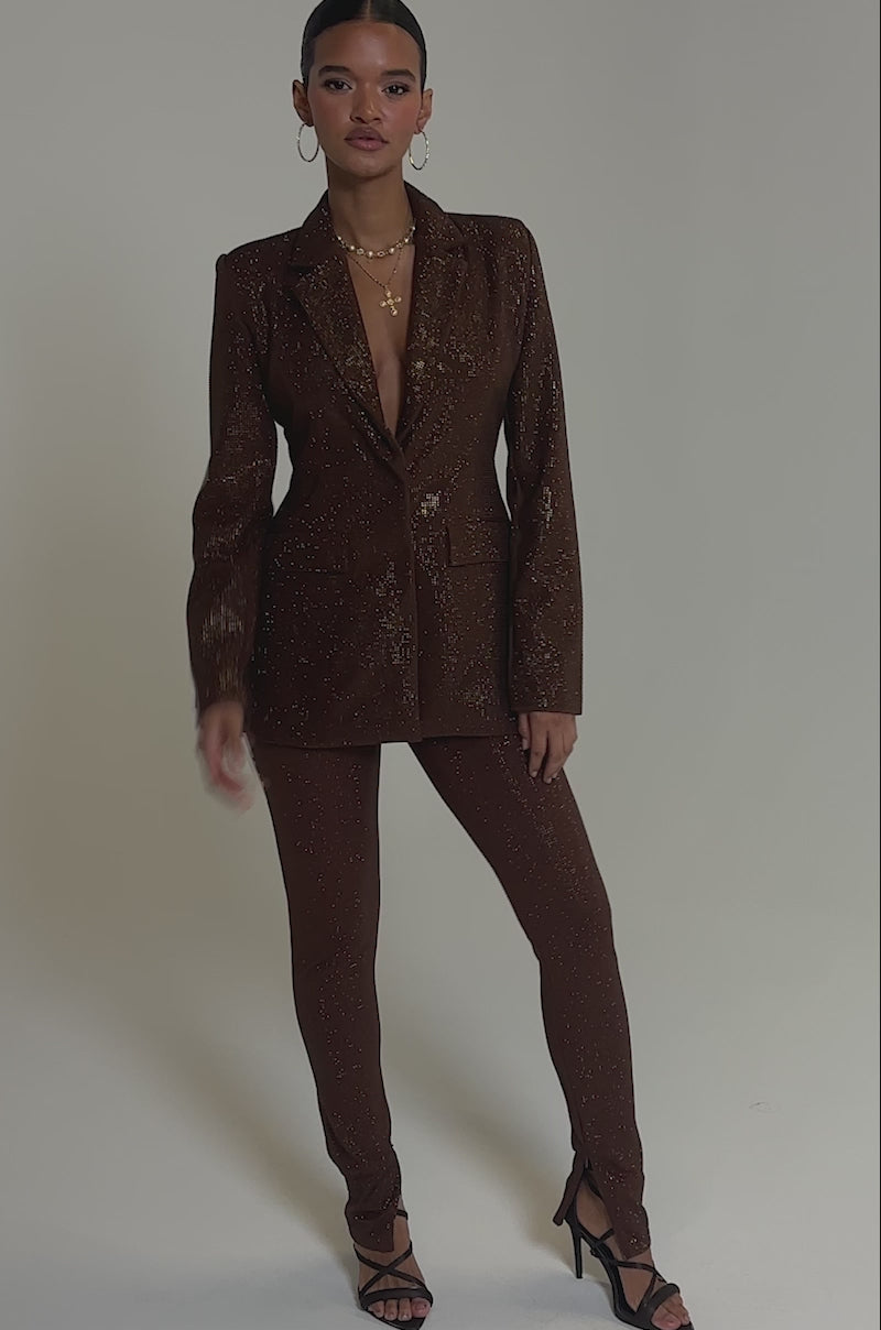 Video of woman modeling the MAKE A TOAST RHINESTONE PANT—high-waisted, brown, skinny-leg pants with rhinestones—paired with a matching long-sleeve blazer and black strappy heels, standing on a simple light background.