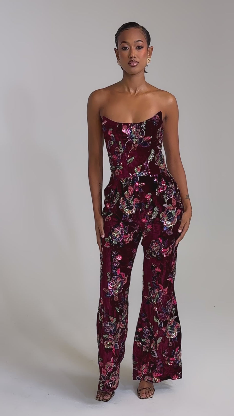 A video of a woman wearing the IN YOUR DREAMS VELVET JUMPSUIT, a strapless burgundy velvet piece with sequin floral embroidery, styled with gold hoop earrings and nude strappy heels against a plain light gray background.