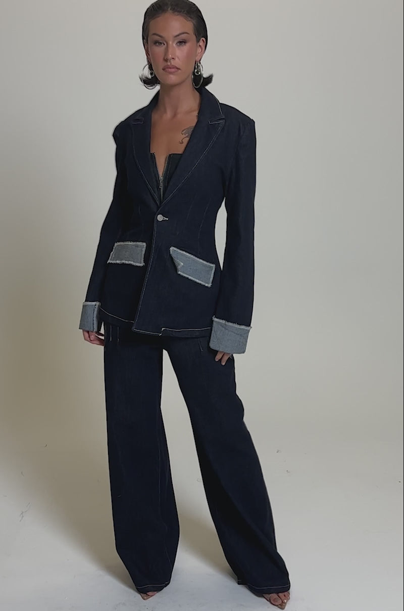 A model poses in the JESSE DENIM BLAZER—a structured dark denim pantsuit with light grey cuff and pocket accents, styled with large hoop earrings and slicked-back hair against a plain background.