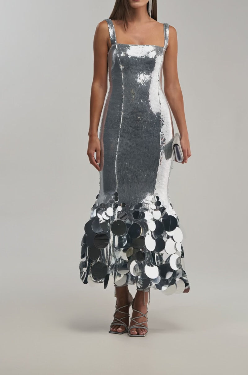 Video of woman modeling the LIFE OF THE PARTY SEQUIN MIDI DRESS IN SILVER, a sleeveless sequin dress with large round silver discs at the hem. She accessorizes with a small clutch and strappy heels against a plain light background.