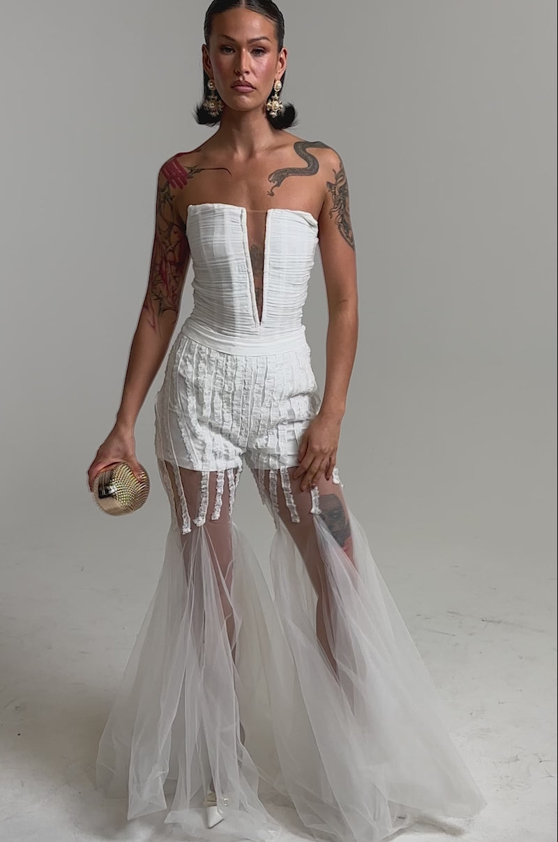 A video of a woman in the "EYE FOR MY EYES ON YOUR MESH FLARE PANT," pairing it with a strapless white V-neck dress, large gold earrings, and a small textured clutch against a plain light background.