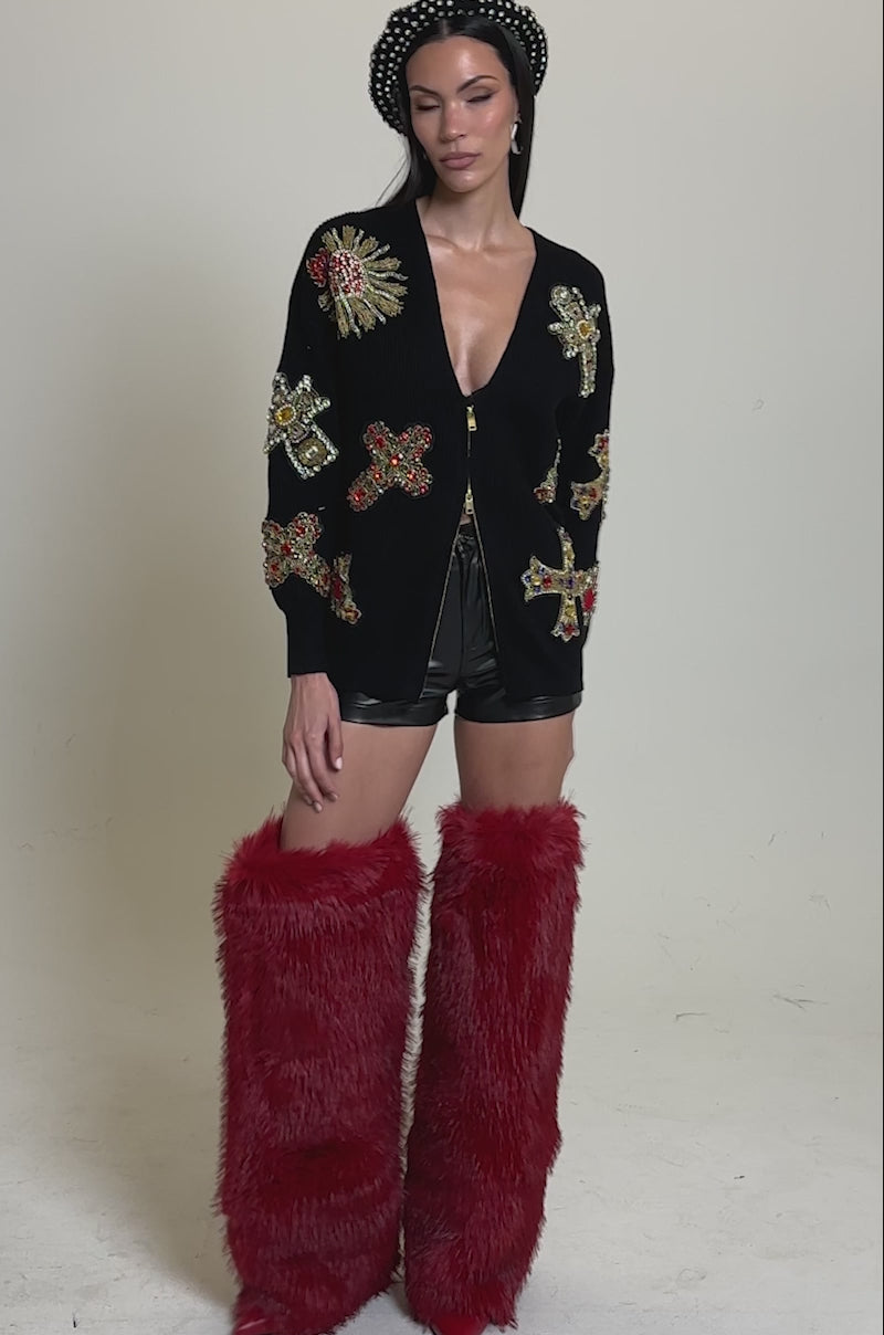 A video of a woman wearing the FEELING EXTRA EMBELLISHED SWEATER with colorful embroidered patches, black shorts, tall red furry boots, and a black beaded headband, standing against a plain background.