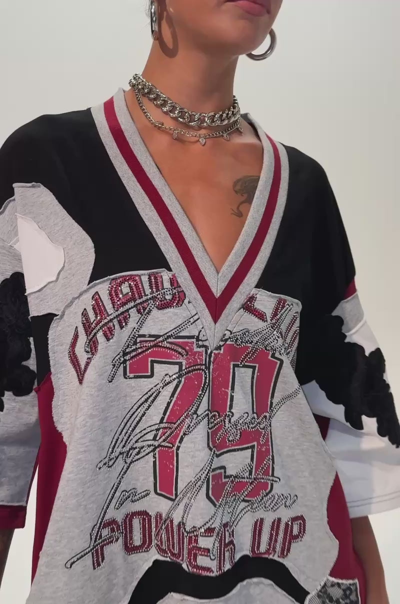 Video of a woman modeling the AFTER HOURS GRAPHIC MINI DRESS, a woman showcases a sporty-chic look in an oversized varsity-inspired silhouette. The dress features striking graphic prints—including "79," "POWER UP," large black leaves, and bold stripes in red, black, white, and gray—doubling as a statement jersey. 