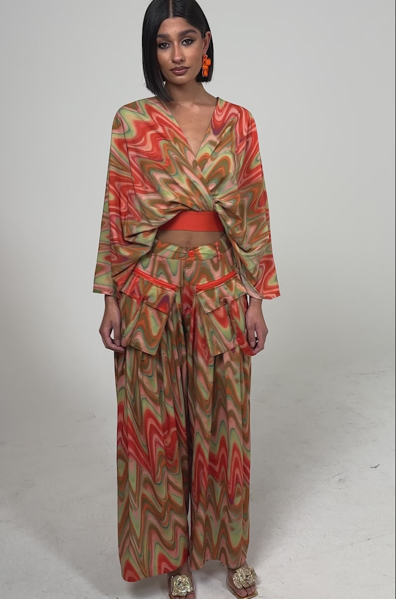 A video of a woman in the DRIFTING AWAY BLOUSE—an abstract swirl wrap top—in sunset hues with wide-leg pants, facing forward against a plain background.