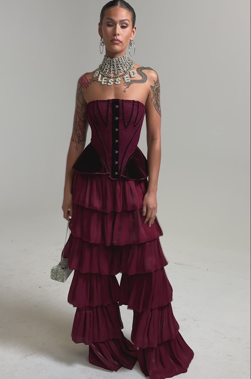 Video of woman modeling the STAR OF THE NIGHT FLOUNCY PANT IN BURGUNDY, featuring high-rise, tiered ruffle wide legs. She pairs it with a strapless burgundy corset, bold statement necklace, and show off arm tattoos against a plain background.