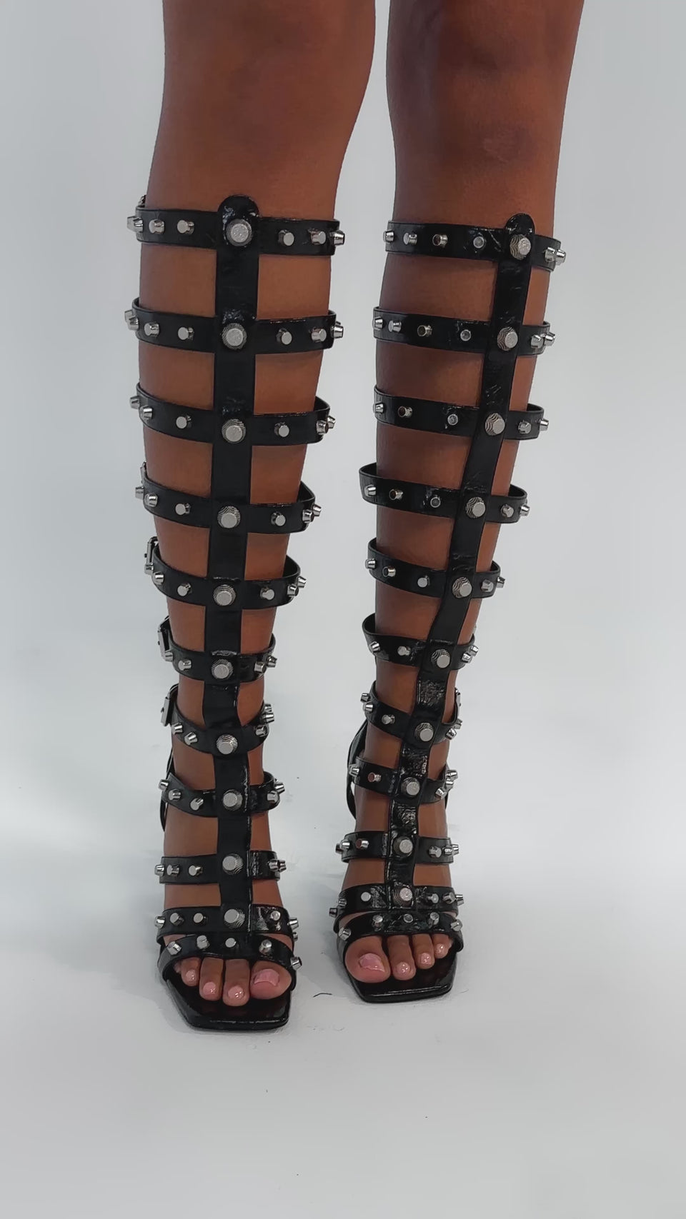 Video of woman modeling the AKIRA GOLD HARDWARE EMBELLISHED BLACK GLADIATOR SANDAL—knee-high, open-toe, with multiple straps, gold hardware, and stiletto heels—against a light gray background.