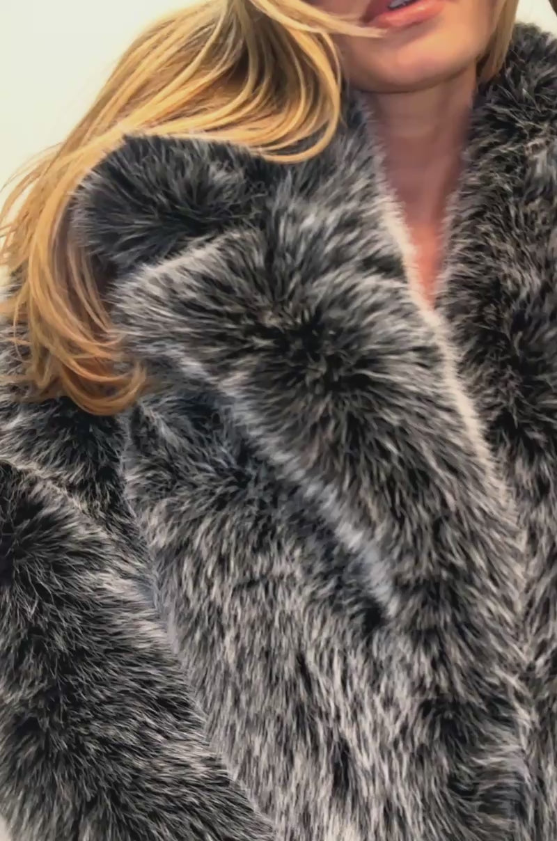 Video of a woman modeling the NO LOOKING BACK FAUX FUR IN GREY, this individual pairs the oversized gray jacket with a black dress, thigh-high stockings, and oversized black sunglasses.