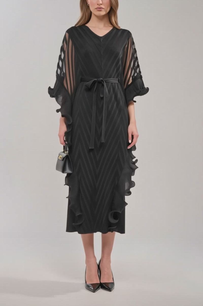 Video of woman modeling the THE NIGHT GOES ON RUFFLE MAXI DRESS IN BLACK, featuring sheer striped sleeves, ruffled edges, a belted waist, and black high heels. She holds a small black handbag and wears her hair loose—ideal for formal occasions.