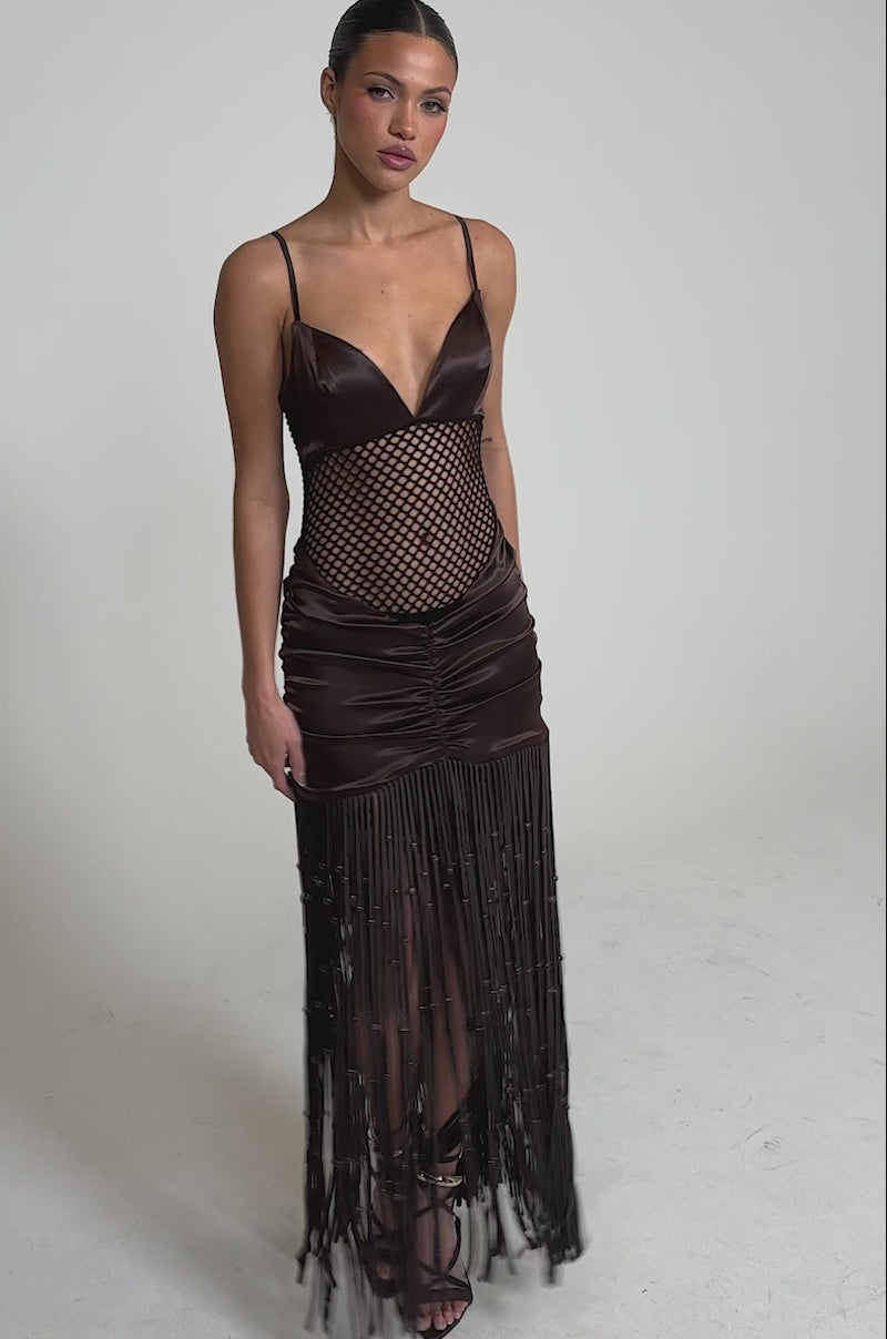 Video of woman modeling the MIDNIGHT MOCHA MAXI DRESS, a sleeveless satin maxi with a deep V-neck, fishnet midsection, and long fringe accents, styled with strappy high-heeled sandals.