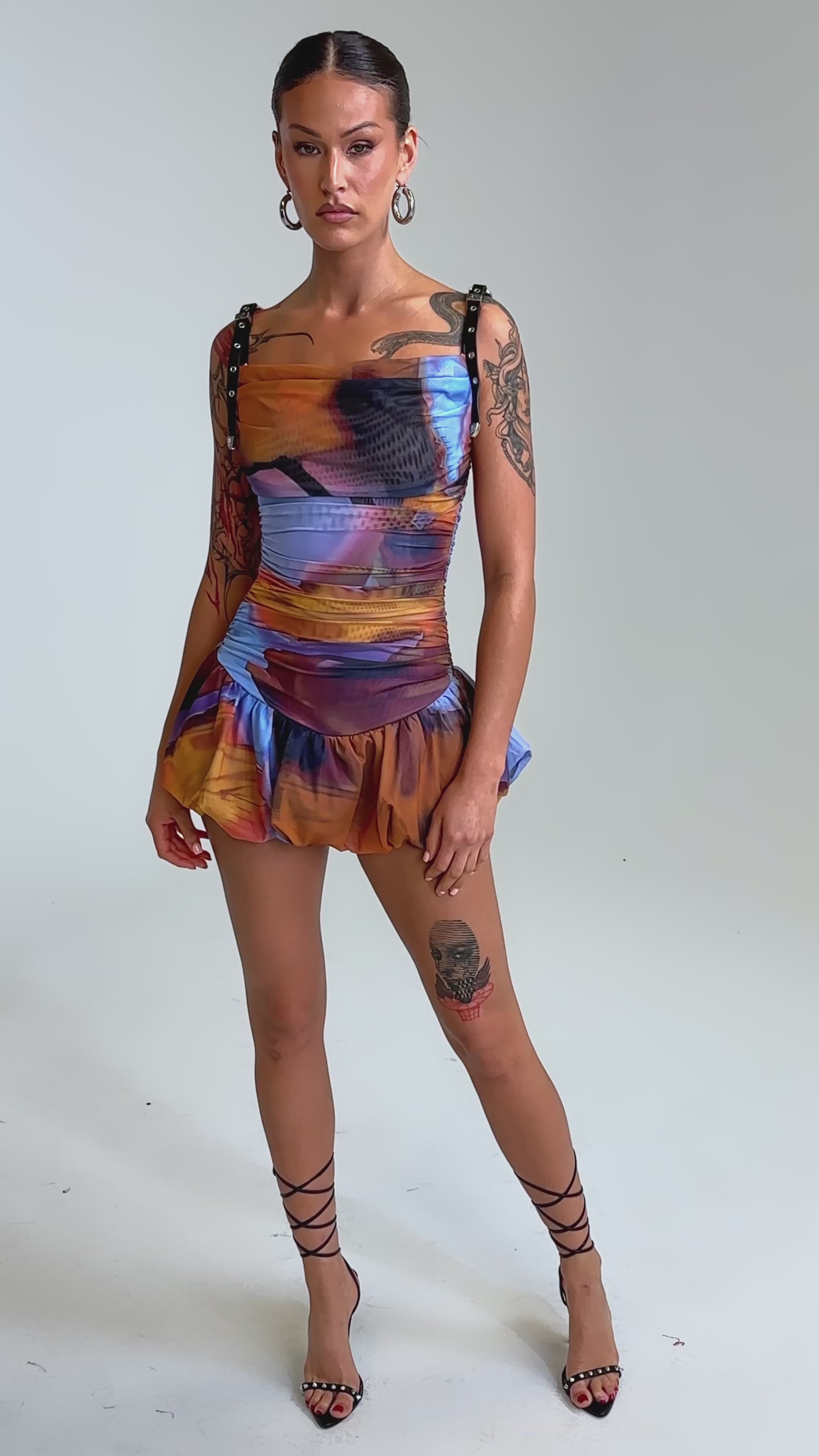 Video of woman modeling the IN A MOOD PRINTED MINI DRESS IN PURPLE—a colorful, form-fitting mini dress with black straps, cowl neck, ruffled hem, and bold blue, orange, and purple tie-dye print—against a light background.