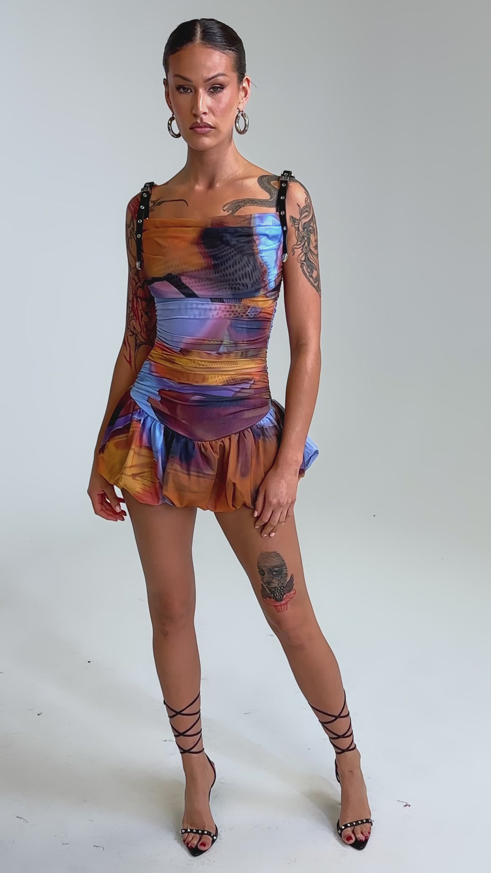 Video of woman modeling the IN A MOOD PRINTED MINI DRESS IN PURPLE—a colorful, form-fitting mini dress with black straps, cowl neck, ruffled hem, and bold blue, orange, and purple tie-dye print—against a light background.