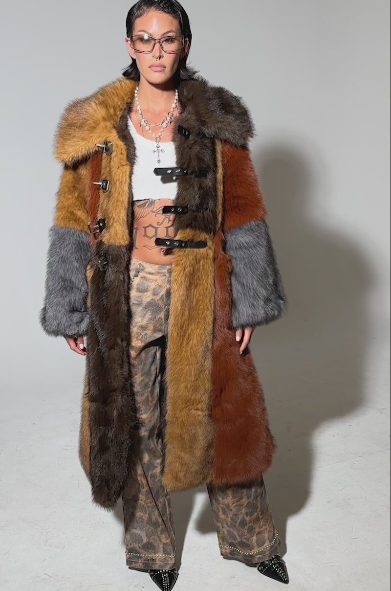 Video of woman modeling the WILD CARD PATCHWORK FAUX FUR coat in multicolored brown, orange, and gray, paired with animal print pants, sunglasses, and black shoes, standing against a plain background