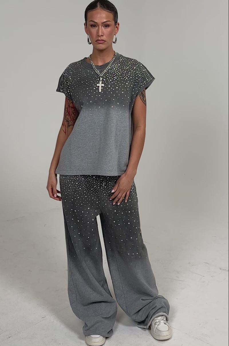 Video of woman modeling the NIGHT SKY RHINESTONE TEE, featuring a lightweight gray knit and small silver studs, paired with matching pants, gold hoop earrings and necklace, and slicked-back hair.