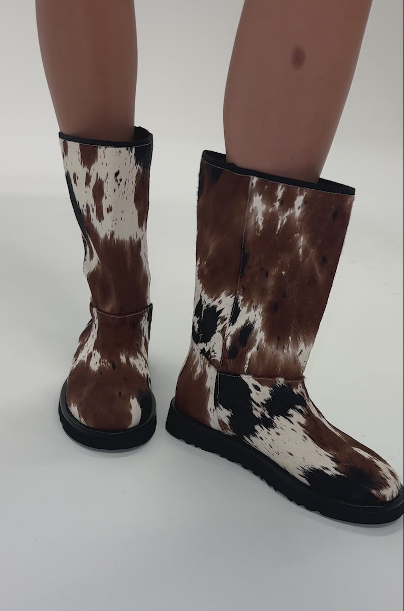 Video of woman modeling the AZALEA WANG MARLEIGH COW FLAT BOOTIE, made from genuine calf hair with brown, white, and black cow print patterns, standing on a light grey background.