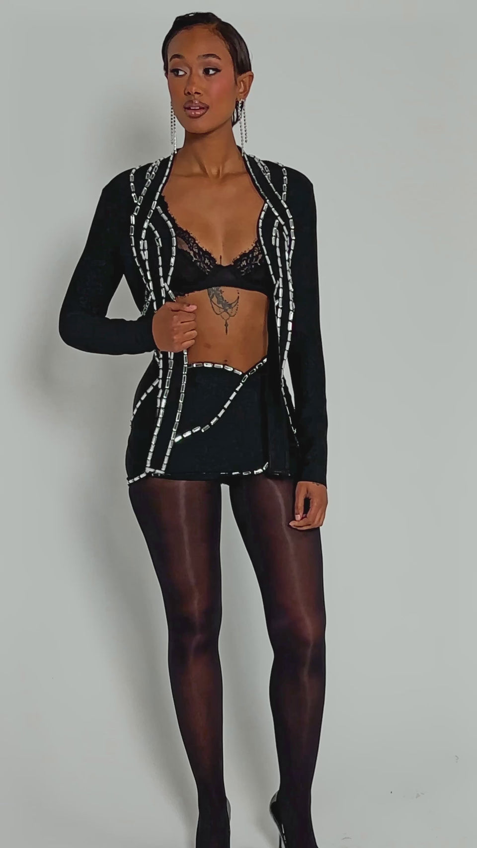 Video of woman modeling the TOO HOT FOR THIS RHINESTONE MINI SKIRT—with a high-rise fit, black fabric, and shiny silver trim—paired with sheer black tights. Photo is cropped from midriff to upper thighs, hands at sides.