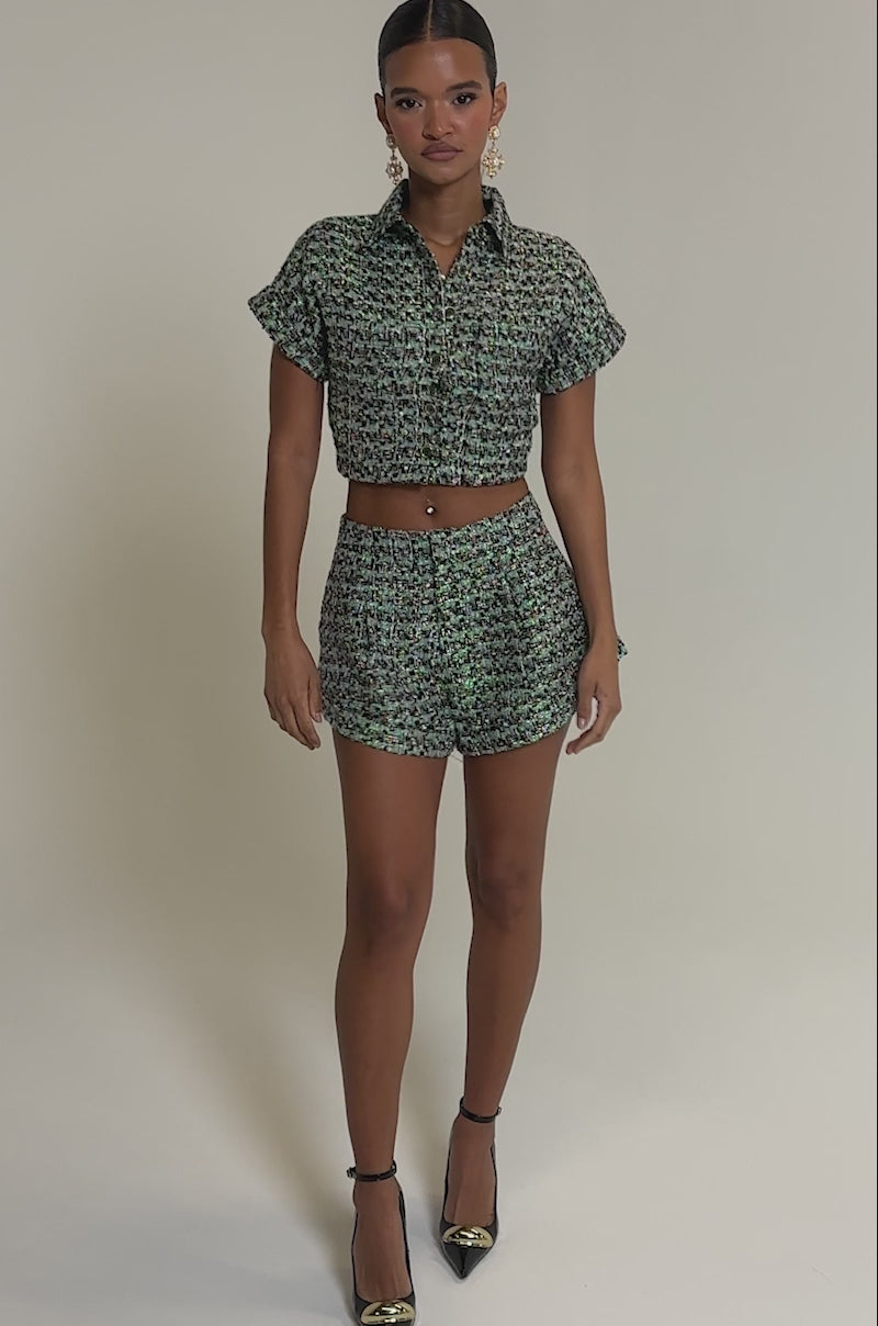 Video of woman modeling the BRIGETTE TWEED BUTTON DOWN as a cropped, textured, multicolored top with matching high-waisted shorts, accessorized with dangling earrings and a belly button piercing against a neutral background.