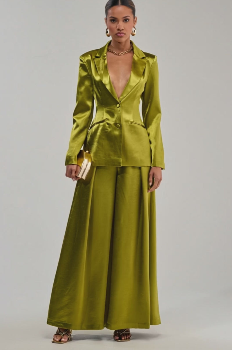 Video of woman modeling the LUCKY ONE SATIN BLAZER and matching green suit, featuring a deep V-neckline. She accessorizes with gold hoop earrings, a chunky necklace, and a round gold clutch—an ideal evening look for special occasions.
