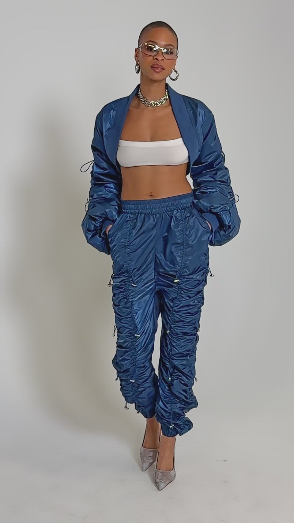 Video of woman modeling the NOT TIME FOR GAMES RUCHED JOGGER IN NAVY with a white bandeau top, blue ruched bomber, hoop earrings, a silver necklace, and high heels