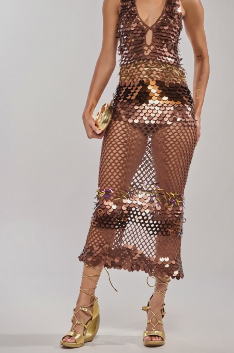 Video of woman modeling the POWER MOVE SEQUIN CROCHET HALTER MAXI DRESS with a deep neckline and mesh skirt holding a gold clutch. She completes her vacation night look with gold strappy heels and styles her curly hair in a high ponytail.