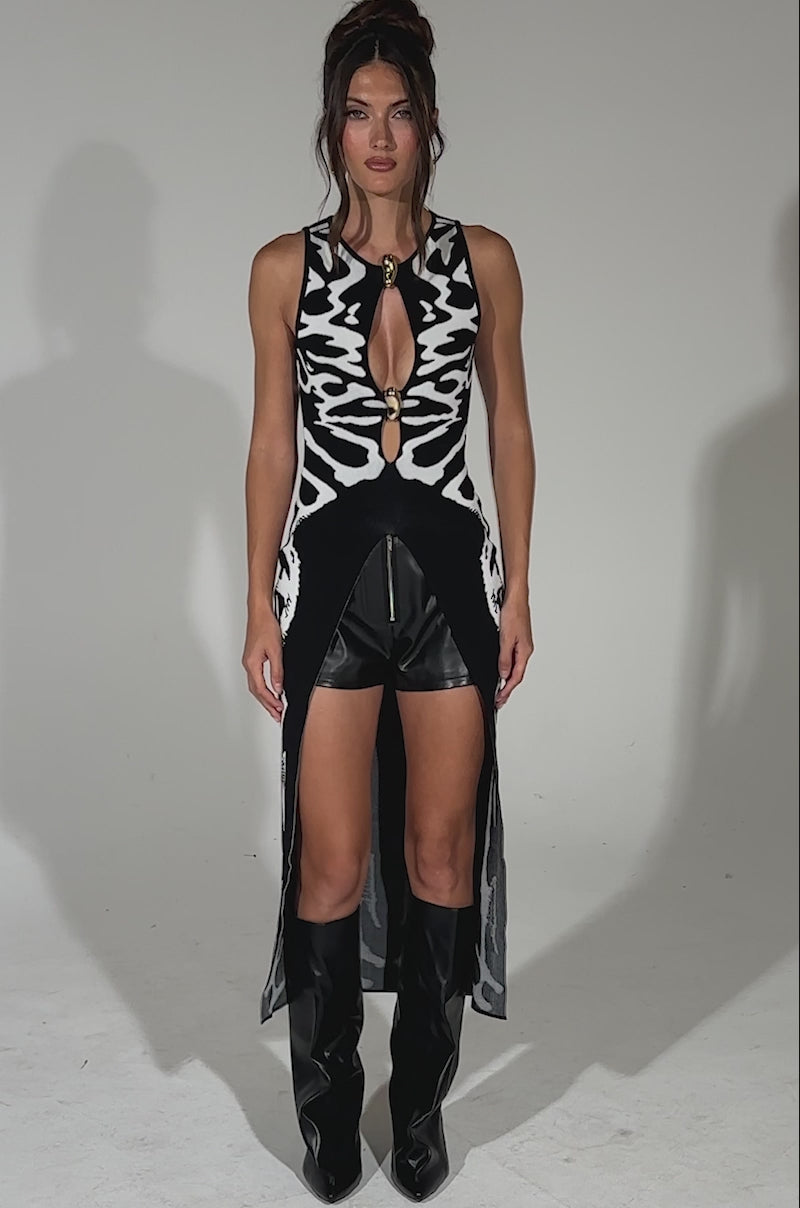 A video of a woman posing against a plain backdrop in the INCOGNITO SWEATER—an abstract black and white knit with a front cutout—paired with black leather shorts, tall black boots, and her hair styled up with soft, face-framing strands.